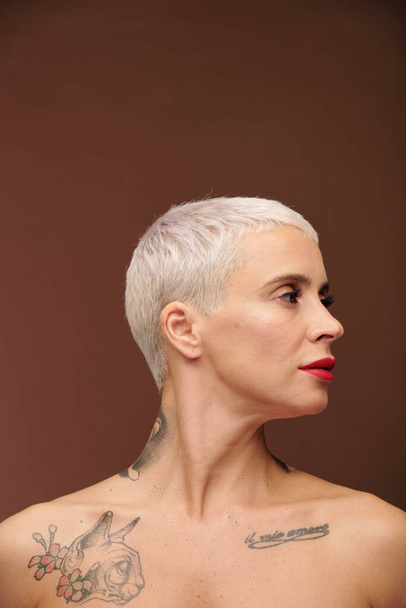 Чоловіча жінка з коротким світлим волоссям, татуювання на плечах, шиї та грудях і червона помада на губах, що тримає голову відвернутою в ізоляції
 - Фото, зображення