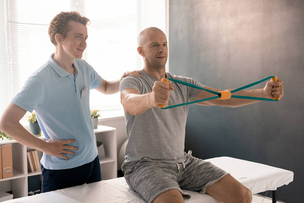 Bald man stretching resistance band during exercise for arms while sitting on medical couch, his physiotherapist standing near by and helping him - Foto, immagini