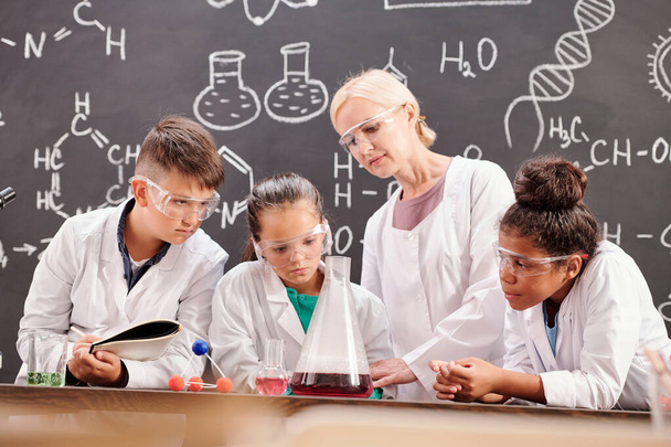 Αρκετοί μαθητές της δευτεροβάθμιας εκπαίδευσης παρακολουθούν χημικό πείραμα που επιδεικνύεται από το δάσκαλό τους της χημείας από το γραφείο στο μάθημα - Φωτογραφία, εικόνα