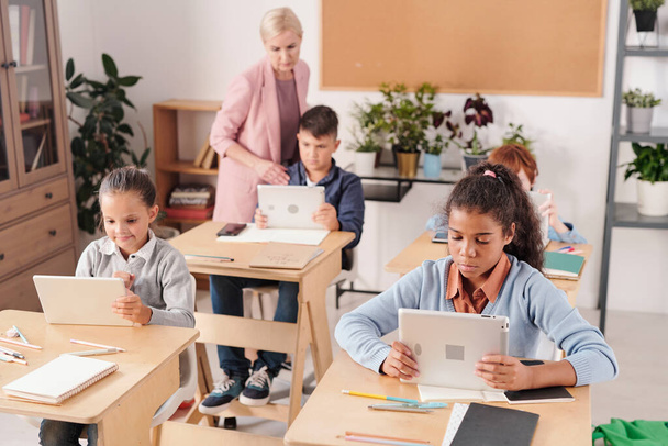 Grupa współczesnych uczniów z tabliczkami siedzącymi przy biurkach w dwóch rzędach i pracującymi indywidualnie, podczas gdy nauczyciel konsultuje się z jednym z nich - Zdjęcie, obraz