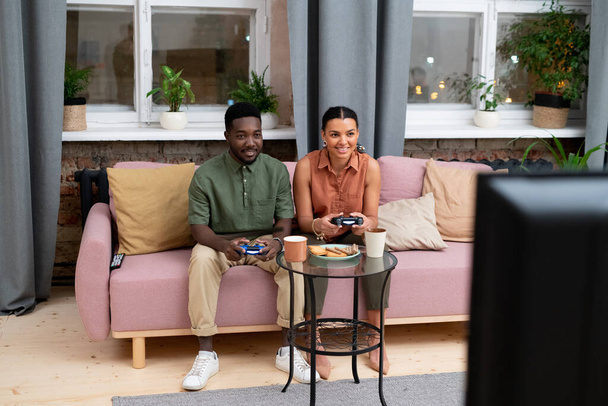 Счастливый подросток и девочка африканской национальности сидят на диване напротив окна в гостиной и играют в видеоигры перед телевизором - Фото, изображение