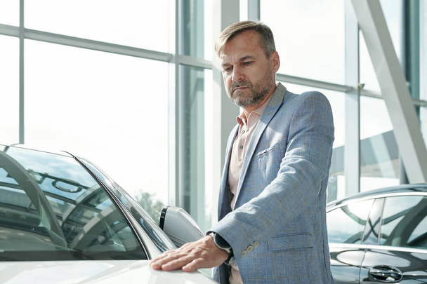 Contemporary mature man in smart casualwear keeping hand on front of new car of white color and looking at it while choosing vehicle - 写真・画像
