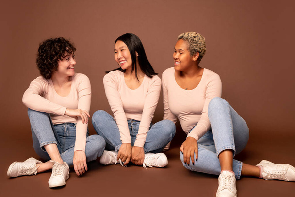 Grupa młodych kobiet o różnym pochodzeniu etnicznym siedzących na podłodze na brązowym tle, patrzących na siebie i rozmawiających - Zdjęcie, obraz