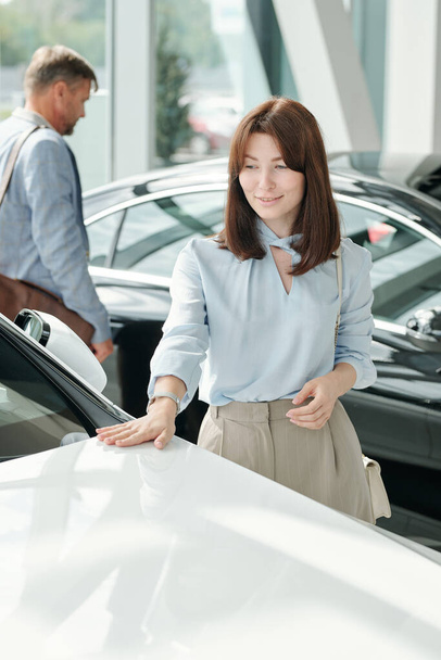 Szczęśliwy młody elegancki brunetka kobieta w smart casualwear dotykając przedniej części nowego luksusowego samochodu w białym kolorze w dużym nowoczesnym centrum auto - Zdjęcie, obraz