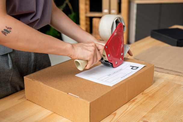 Hands of young female manager of online shop with cellotape gun dispenser sealing cardboard box with packed order of client by table - Photo, Image