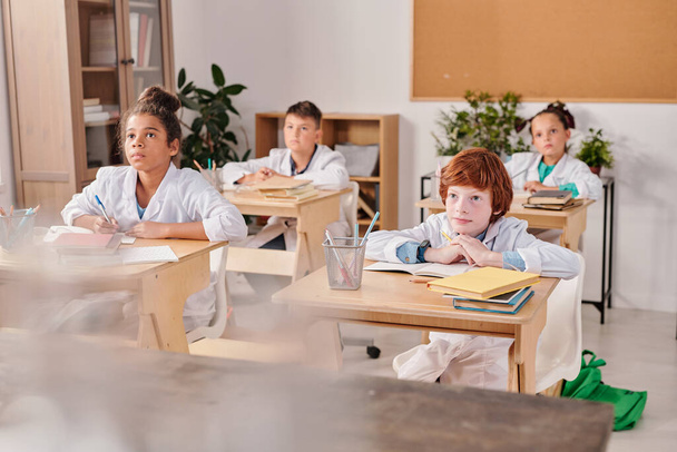 Několik žáků základních škol v bílých pláštích sedí ve dvou řadách u psacích stolů na hodině chemie a poslouchají, o čem jejich učitel mluví - Fotografie, Obrázek