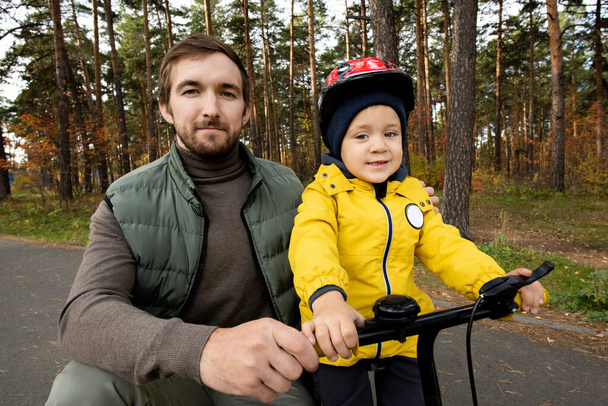 Joven padre feliz en ropa casual en cuclillas por su lindo hijo pequeño en casco de seguridad sosteniendo por asas de bicicleta de equilibrio, mientras que durante el frío en el parque - Foto, imagen