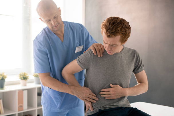 Больной человек показывает врачу, где болит и трогает спину, сидя на медицинском диване во время реабилитационной тренировки в клиниках - Фото, изображение