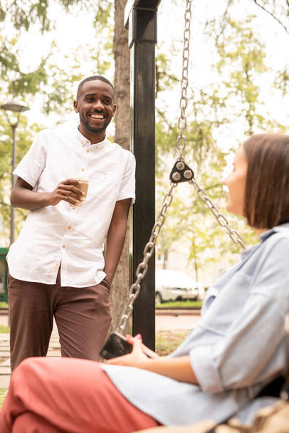 Fröhlicher afrikanischer Kerl mit Getränk steht vor einer jungen Frau, die es sich auf einer Schaukel gemütlich macht, während sie an einem Sommertag im Park mit ihr spricht - Foto, Bild