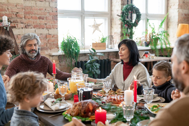 Tres generaciones de familia numerosa sentada junto a la mesa festiva servida y cogida de la mano mientras rezaba antes de la cena de Navidad en casa - Foto, imagen