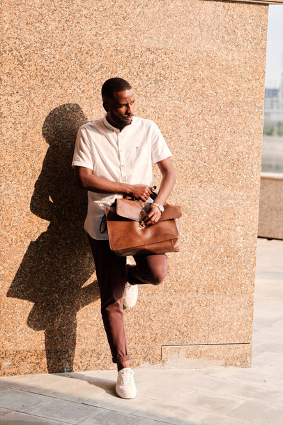 Young African businessman or agent in smart casualwear holding brown leather handbag on his knee while leaning against wall of building - Photo, Image
