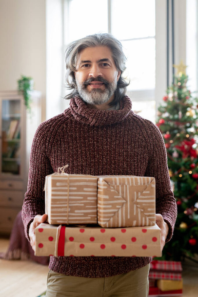 Gelukkig volwassen man in casualwear houden verpakt cadeautjes voor zijn vrouw en kleinkinderen terwijl staan tegen versierde kerstboom - Foto, afbeelding