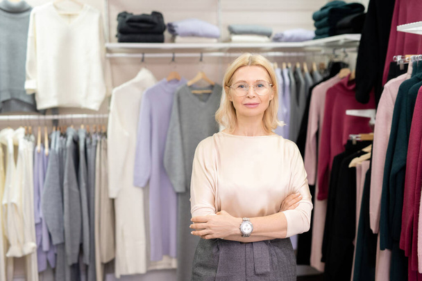 Blond mature female shop assistant in casualwear and eyeglasses crossing arms by chest while standing in clothing department against racks - Photo, image