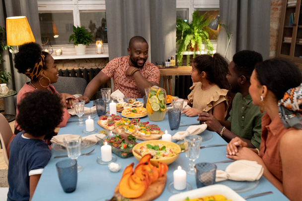 Glücklicher junger afrikanischer Mann schaut seine süße Tochter an und spricht mit ihr bei einem festlichen Tisch während des Weihnachts- oder Thanksgiving-Abendessens - Foto, Bild