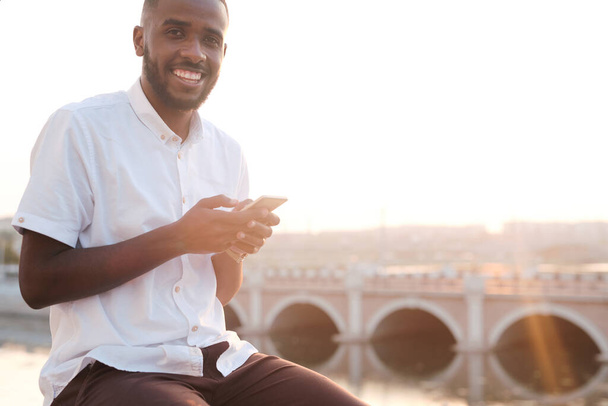 Young smiling African businessman in white shirt and brown pants using smartphone while sitting in front of camera in urban environment - Valokuva, kuva