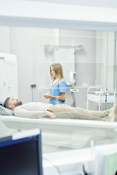Joven doctora rubia con uniforme médico de pie junto a un paciente barbudo que yace en la litera durante un examen ultrasonido en clínicas - Foto, Imagen