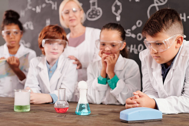 Colegiales serios e inteligentes en gafas blancas y gafas protectoras que observan diferentes reacciones en tubos de vidrio con compuestos químicos en la lección - Foto, Imagen