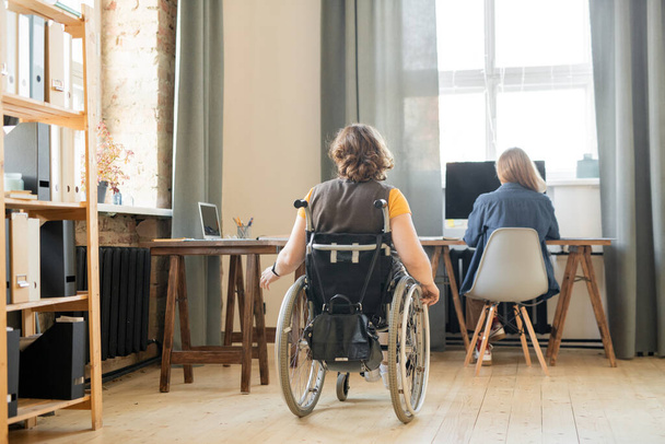 Rückenansicht einer jungen behinderten Frau im Rollstuhl, die sich an einem der Tische im großen modernen Büro bewegt, wo ihr Kollege arbeitet - Foto, Bild