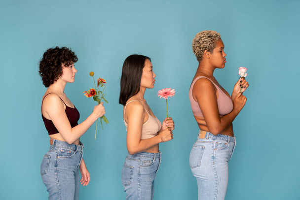 Πλάγια άποψη τριών νεαρών διαπολιτισμικών γυναικών σε πάγκους και τζιν που κρατούν φρέσκα λουλούδια μπροστά τους ενώ στέκονται στη σειρά - Φωτογραφία, εικόνα