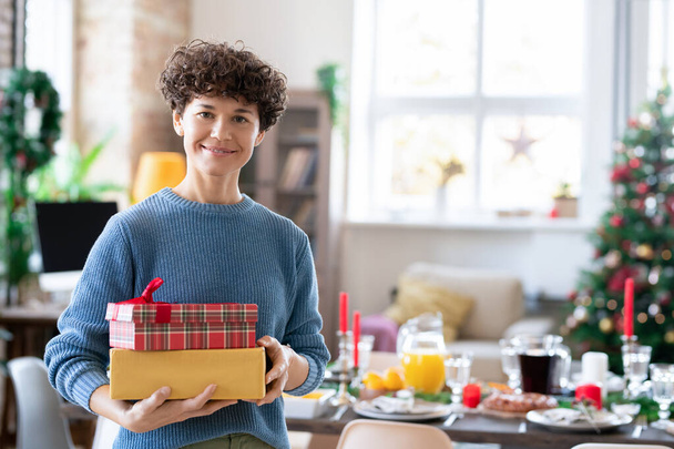 młody uśmiech kręcone brunetka kobieta gospodarstwa pudełka z prezentami dla jej rodziny stojąc przed urządzone choinki i służył stół - Zdjęcie, obraz