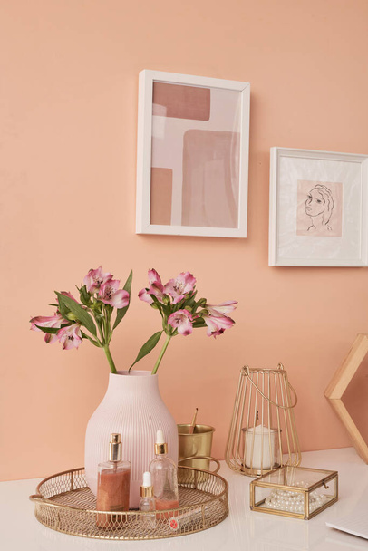 Duas imagens em quadros na parede rosa por mesa com flores em vaso, produtos de cuidados de beleza, vela, colar de pérolas, balde de ouro e outras coisas - Foto, Imagem