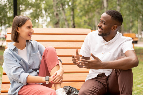 Happy young intercultural couple in casualwear sitting on wooden bench in park or square, chatting and enjoying summer day at leisure - Photo, Image