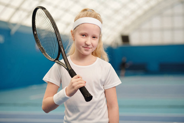 Mooi blond klein meisje in witte activewear houden tennis racket door rechter schouder terwijl staan in de voorkant van de camera in stadion omgeving - Foto, afbeelding