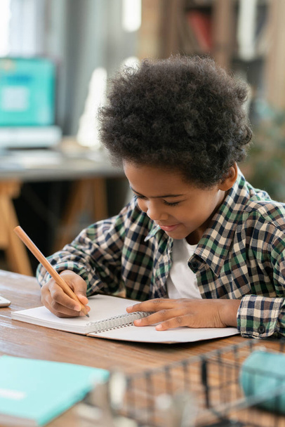 Cute elementary schoolboy of African ethnicity making notes in copybook while doing homework by table in living-room against computer - Foto, Imagem