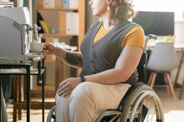 Tekerlekli sandalyedeki genç iş kadını kahve makinesinin önünde oturuyor ve günün ortasında mola verirken sıcak bir şeyler hazırlıyor. - Fotoğraf, Görsel