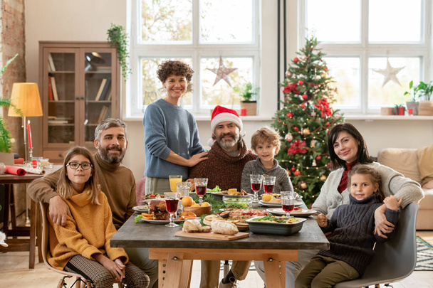 Heureux couples jeunes et matures et trois mignons enfants réunis par table de fête servie pour le dîner de Noël dans le salon contre arbre décoré - Photo, image