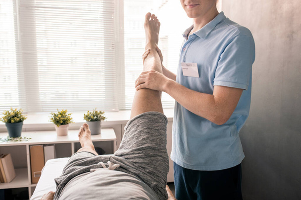 Jonge fysiotherapeut houdt been van de patiënt terwijl hem te helpen met een van de fysieke oefeningen in moderne revalidatiecentrum of klinieken - Foto, afbeelding