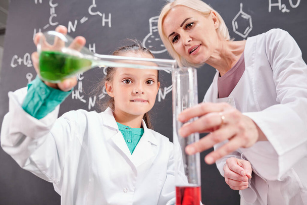 Profesora rubia confiada de química y colegiala en whitecoat mezclando sustancia líquida verde en tubo con roja contra pizarra - Foto, Imagen