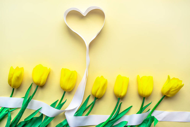 Ηλιόλουστη κίτρινη σύνθεση λουλουδιών. Κίτρινες φρέσκες τουλίπες με καρδιές σε κίτρινο χάρτινο φόντο. Ευχετήρια κάρτα για την Ημέρα του Αγίου Βαλεντίνου, Ημέρα της Μητέρας ή άλλες διακοπές. Επιλεκτική εστίαση. Επίπεδη. Αντιγραφή χώρου. - Φωτογραφία, εικόνα