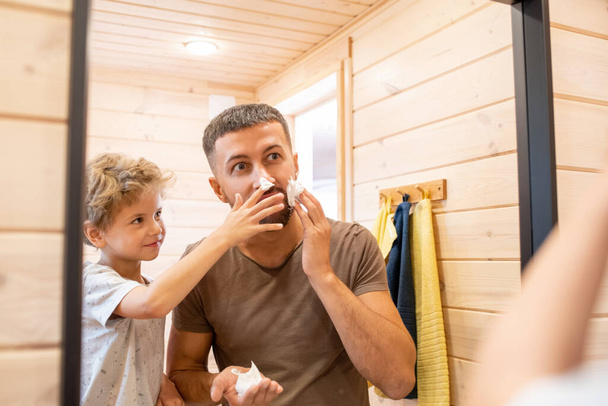 Cute little boy having fun with his father applying foam on face before shaving while both standing in front of mirror in the morning - Foto, immagini