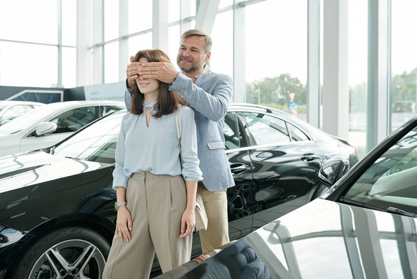 Ευτυχισμένος ώριμος άνδρας που καλύπτει τα μάτια της συζύγου του στέκεται ανάμεσα σε δύο νέα αυτοκίνητα σε μεγάλο σύγχρονο κέντρο αυτοκινήτων, ενώ ο σύζυγός της πρόκειται να αγοράσει ένα - Φωτογραφία, εικόνα