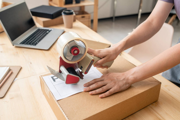 Hands of female manager of online shop with cellotape gun dispenser sealing cardboard box with packed order of client by her workplace - Photo, Image