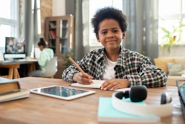 Adorable happy schoolboy of African ethnicity looking at you while making notes in copybook by table against his sister sitting by computer - Foto, immagini