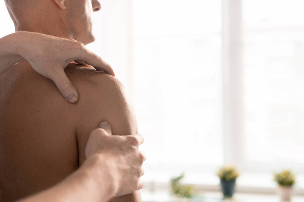 Ruce profesionálního maséra nebo fyzioterapeuta masírujícího ramena dospělého muže bez trička sedícího před ním na klinikách - Fotografie, Obrázek