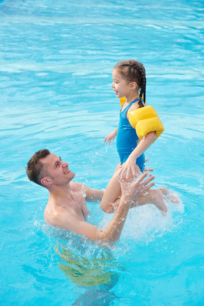 Happy young man playing with his cute little daughter in swimming pool in front of camera while spending weekend together in spa center - Foto, afbeelding