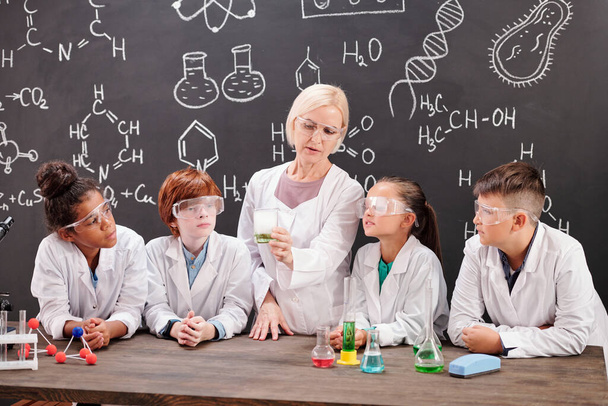 Giovane insegnante biondo di chimica in whitecoat mostrando gruppo di simpatici alunni miscela di due sostanze chimiche mentre discutono la reazione - Foto, immagini