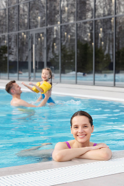 Szczęśliwa młoda brunetka patrzy na ciebie z uśmiechem podczas relaksu w basenie przeciwko mężowi i córce bawiących się w wodzie - Zdjęcie, obraz