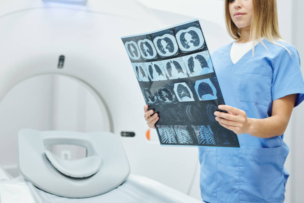 Jovem radiologista feminina em uniforme azul segurando imagem de raio-X da cabeça do paciente e do cérebro contra a máquina de ultra som no consultório médico moderno - Foto, Imagem