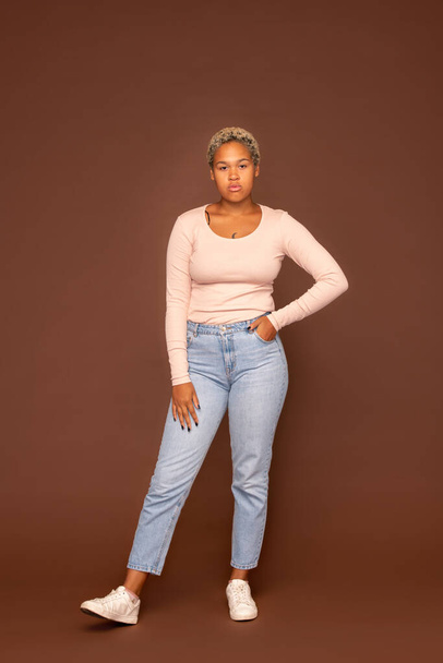 Young serious mixed-race female in blue jeans and white pullover keeping her right arm on leg while standing on brown background in isolation - Φωτογραφία, εικόνα