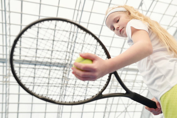 Ernstes junges blondes Mädchen in weißer Aktivkleidung hält Tennisball unter Schläger, während es sich im Stadion auf das Spiel vor der Kamera vorbereitet - Foto, Bild