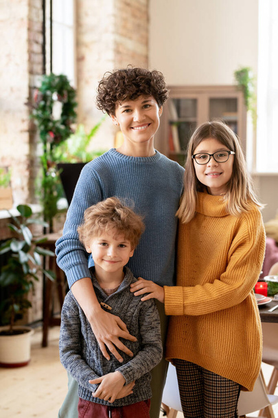 Genç neşeli esmer kadın, gündelik kıyafetleriyle iki tatlı çocuğunu kucaklıyor Noel akşamı kameranın önünde dikilirken. - Fotoğraf, Görsel
