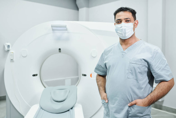 Successful young mixed-race male doctor in protective mask and uniform standing in front of camera against ultra sound equipment - Photo, Image