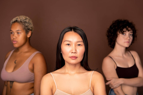 Jovem mulher asiática séria em bege tananabol em pé na frente da câmera contra duas outras fêmeas de várias etnias e olhando para você - Foto, Imagem