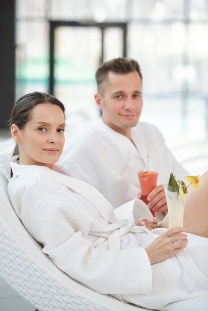Young restful couple in white bathrobes having tropical cocktails while sitting on deckchairs in front of camera in luxurious spa center - Valokuva, kuva