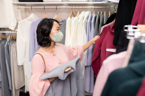 Joven cliente femenino asiático en máscara y blusa inteligente rosa de pie por rack con nuevo surtido de ropa casual y suéter de elección - Foto, imagen