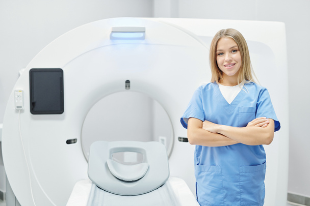 Erfolgreiche junge hübsche Ärztin in blauer Uniform, die die Arme an der Brust kreuzt, während sie vor der Kamera gegen eine Ultraschallmaschine steht - Foto, Bild